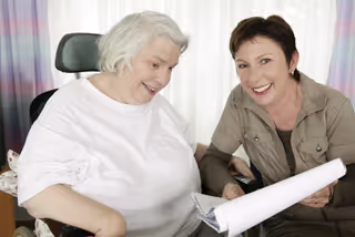 Klientin und Pflegerin sitzend auf einem Stuhl, Blick auf den Pflegeplan